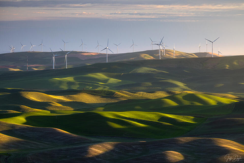 Rolling green hills of the Palouse at sunrise viewed from Steptoe Butte, with shadows across the fields and wind turbines lining a distant ridge beneath clouds