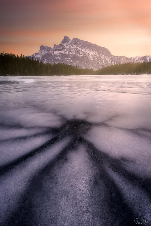 Ice fracture patterns radiating across frozen Two Jack Lake at winter sunset with Mount Rundle rising against a rose sky, Banff National Park