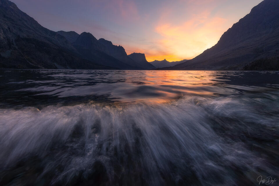Sunset at Saint Mary Lake in Glacier National Park with a soft breaking wave in the foreground and glowing pastel light reflecting across the lake 