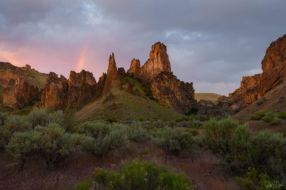 Golden light on rugged rock spires in Leslie Gulch, Oregon, with a faint rainbow, pink and grey clouds, and sagebrush below.