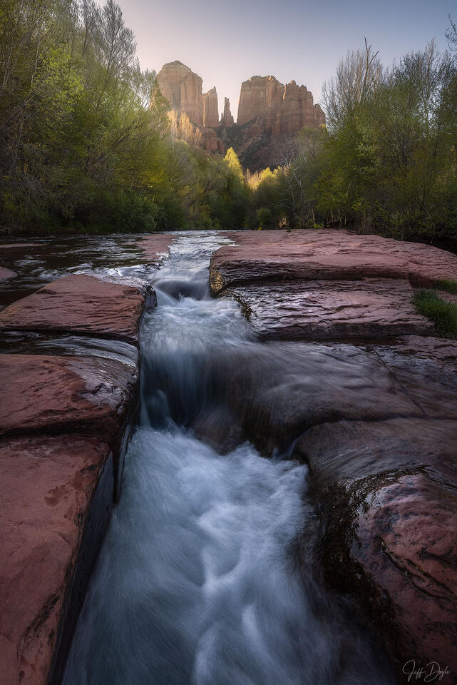 Sedona creek flowing over red rock with Cathedral Rock in the distance at sunrise, smooth water motion and early morning light.