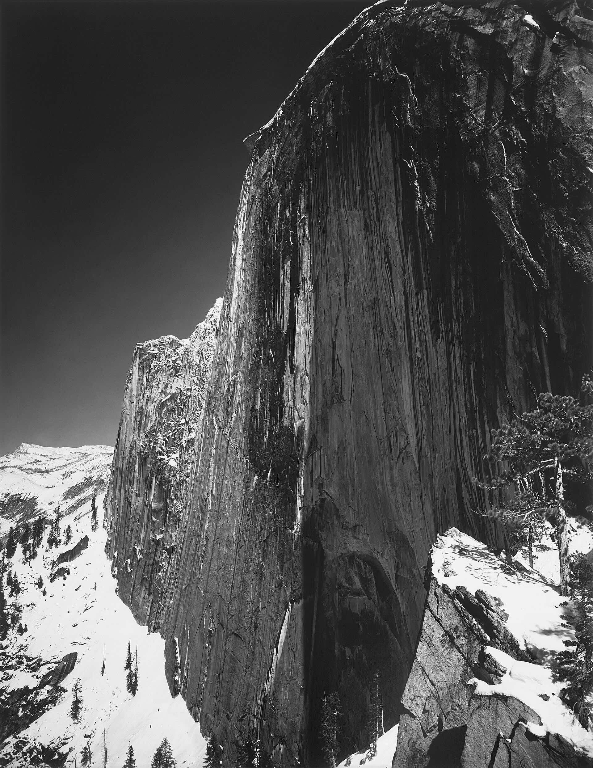 Black-and-white photograph of the massive granite face of El Capitan rising steeply above a snow-covered slope in Yosemite Valley, with dark vertical rock textures contrasting against bright winter snow and a deep, cloudless sky.