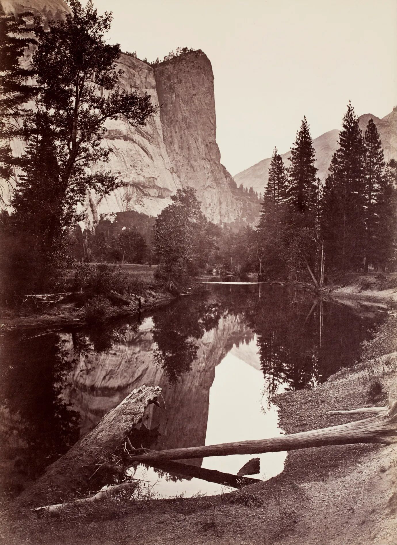 Historic sepia-toned photograph of Yosemite Valley featuring the towering granite face of El Capitan rising above a calm river. Tall pine trees line the water’s edge, and the cliff and forest reflect clearly in the still surface, capturing the quiet grandeur of Yosemite’s landscape.