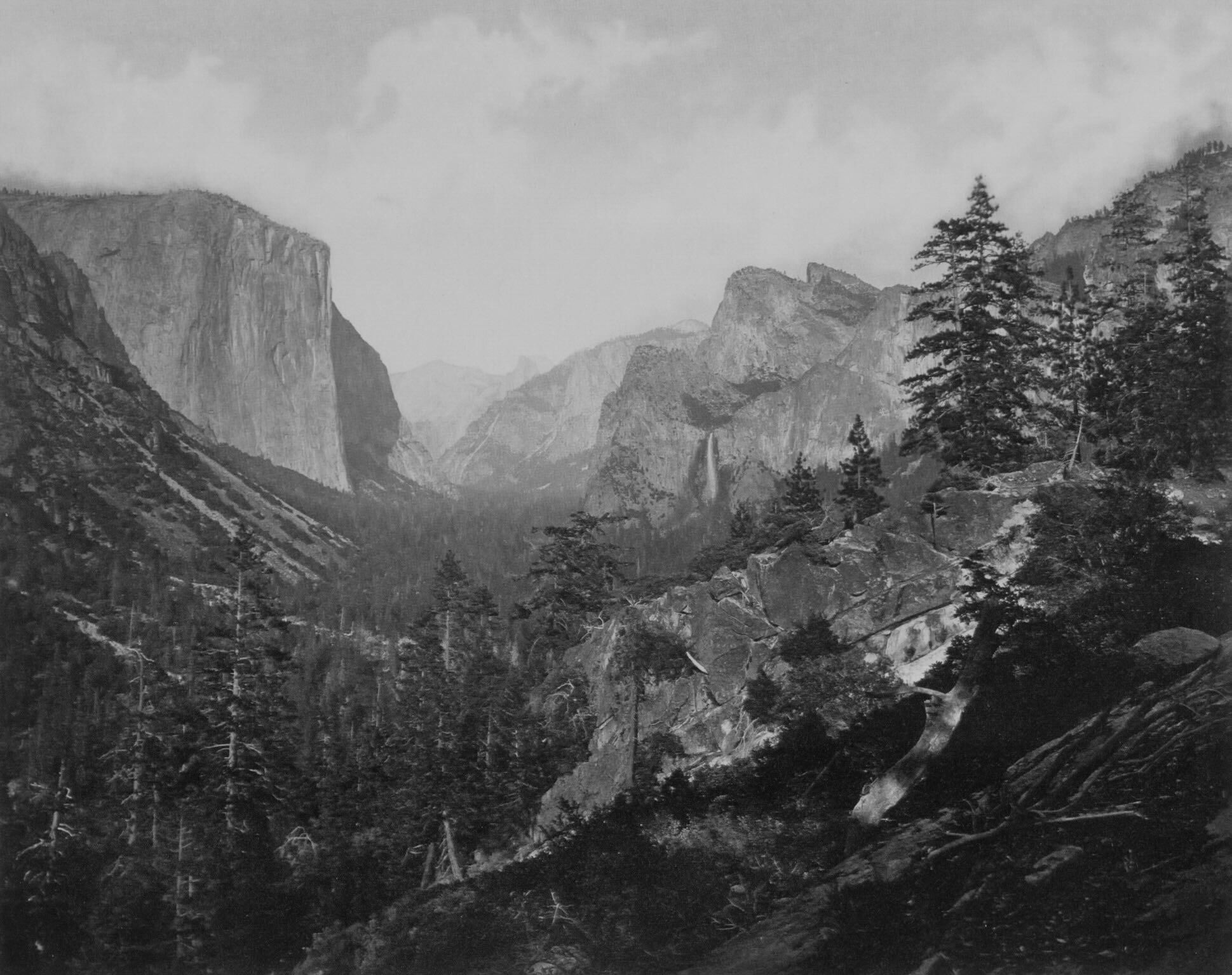 Historic black-and-white photograph of Yosemite Valley with the massive granite face of El Capitan rising on the left and Bridalveil Fall cascading in the distance, surrounded by dense pine forests and rugged granite outcrops in the foreground.