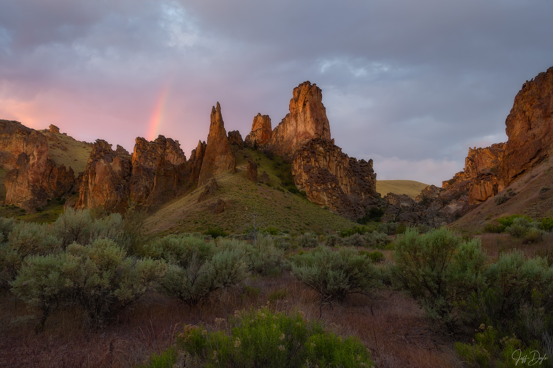 A Quiet Promise Golden light on rugged rock spires in Leslie Gulch, Oregon, with a faint rainbow, pink and grey clouds, and sagebrush below.