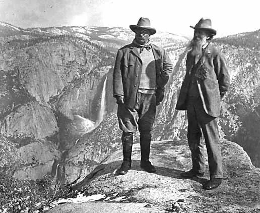 Historic black-and-white photograph of Theodore Roosevelt and John Muir standing on a rocky overlook in Yosemite Valley. Both men wear wide-brimmed hats and outdoor clothing while towering granite cliffs and a distant waterfall rise behind them, symbolizing the early conservation movement in America.