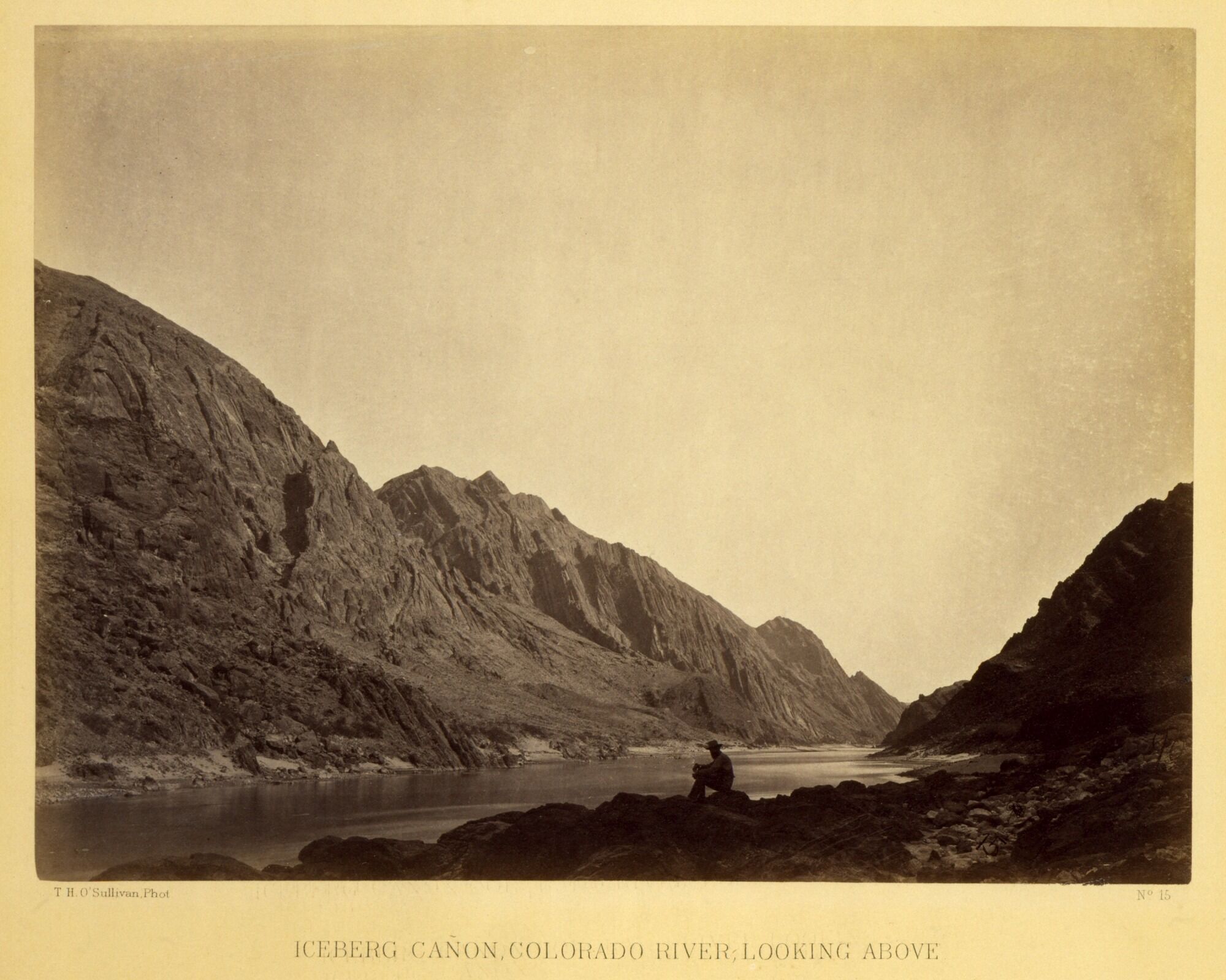 Historic photograph of Iceberg Canyon along the Colorado River by Timothy O’Sullivan, showing steep rock walls, a calm river, and a lone seated figure in the foreground