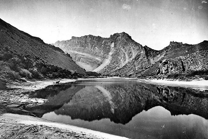 Historic black-and-white photograph of the Green River winding through a rugged canyon landscape, with steep desert mountains rising on both sides. The calm river reflects the jagged peaks and barren slopes above, while sandy banks and sparse vegetation line the foreground, capturing the stark beauty of the American West.